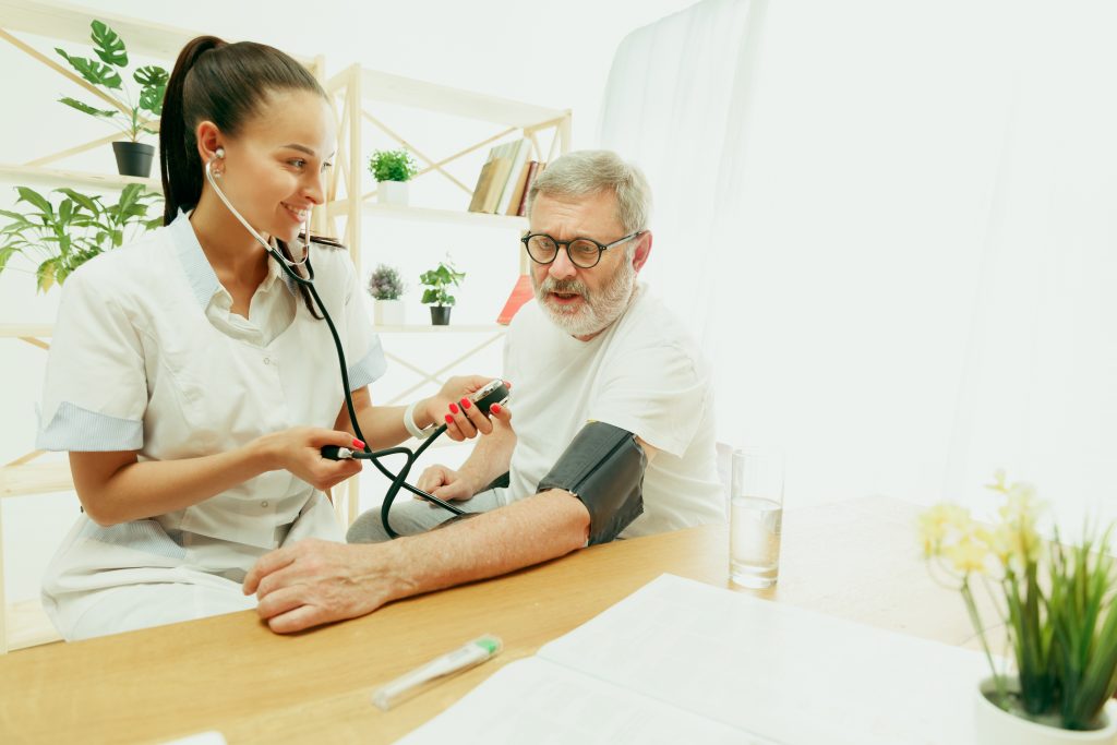 The visiting nurse or health visitor taking care of senior man. Lifestyle portrait at home. Medicine, healthcare and prevention. Girl checking or measuring patient's blood pressure during the visit.