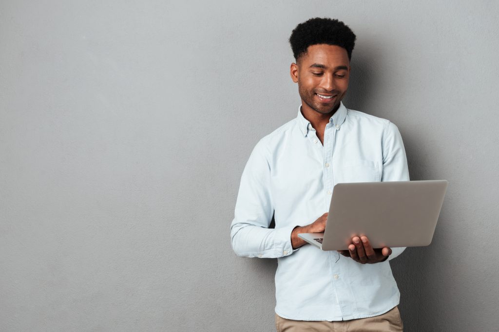 Young smiling african man standing and using laptop computer isolated over gray background